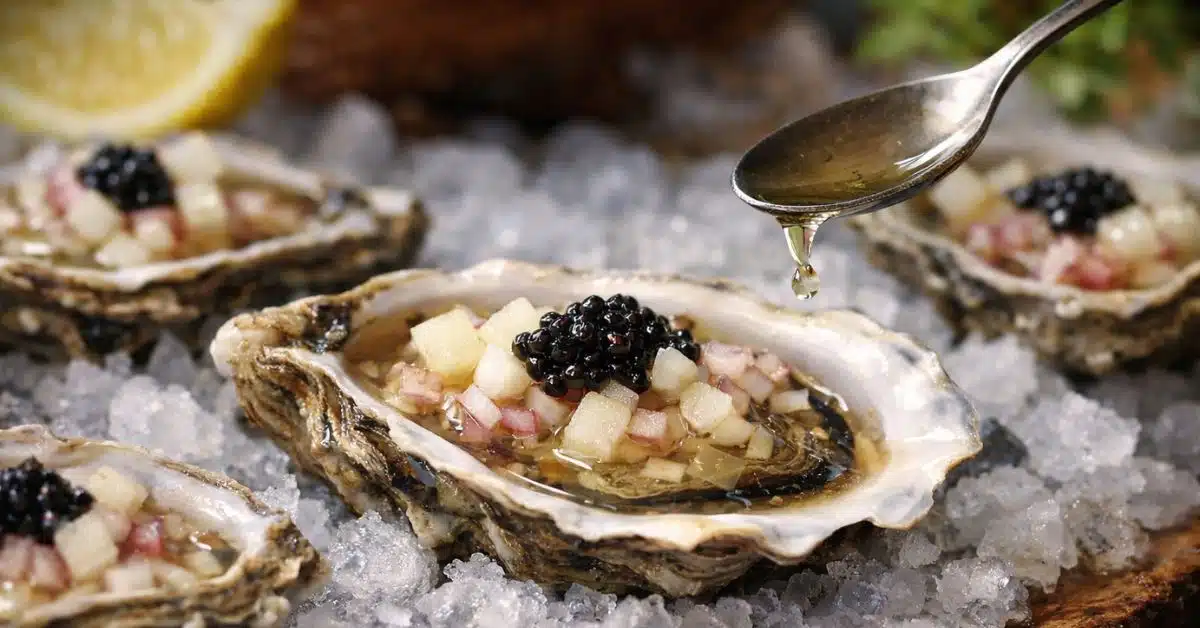 Close-up of freshly shucked Pacific oysters on crushed ice, topped with finely diced pear, minced shallots, and black roe, as champagne vinegar is gently poured over the oyster.